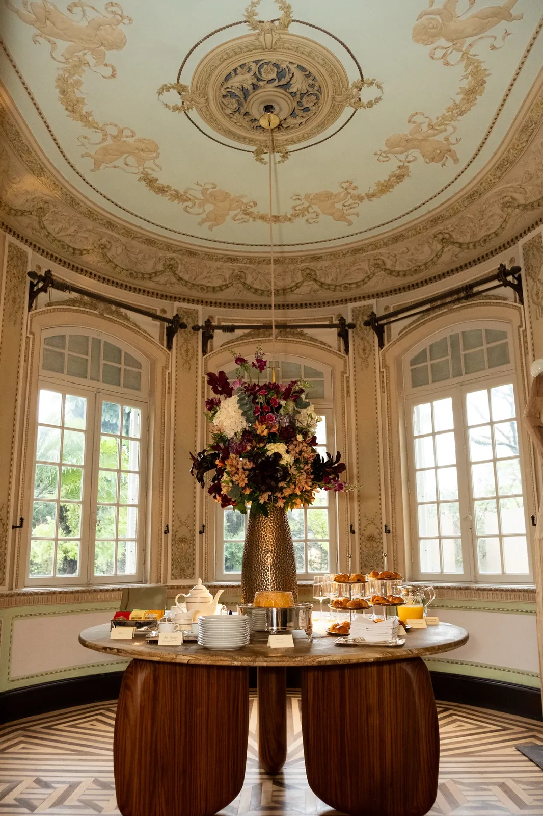 Mesa de café da manhã em sala oval, sob forro decorado com querubins e guirlandas.