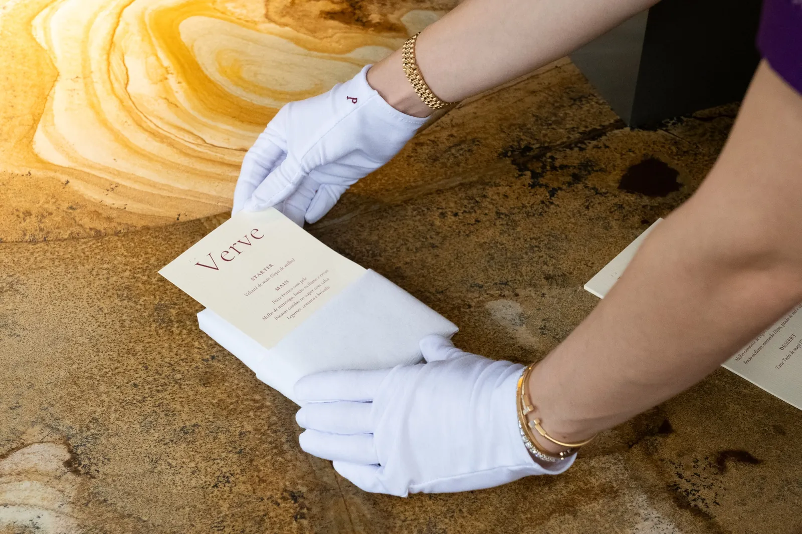 Mãos enluvadas de branco dobram o guardanapo sobre o menu impresso da La Verve.