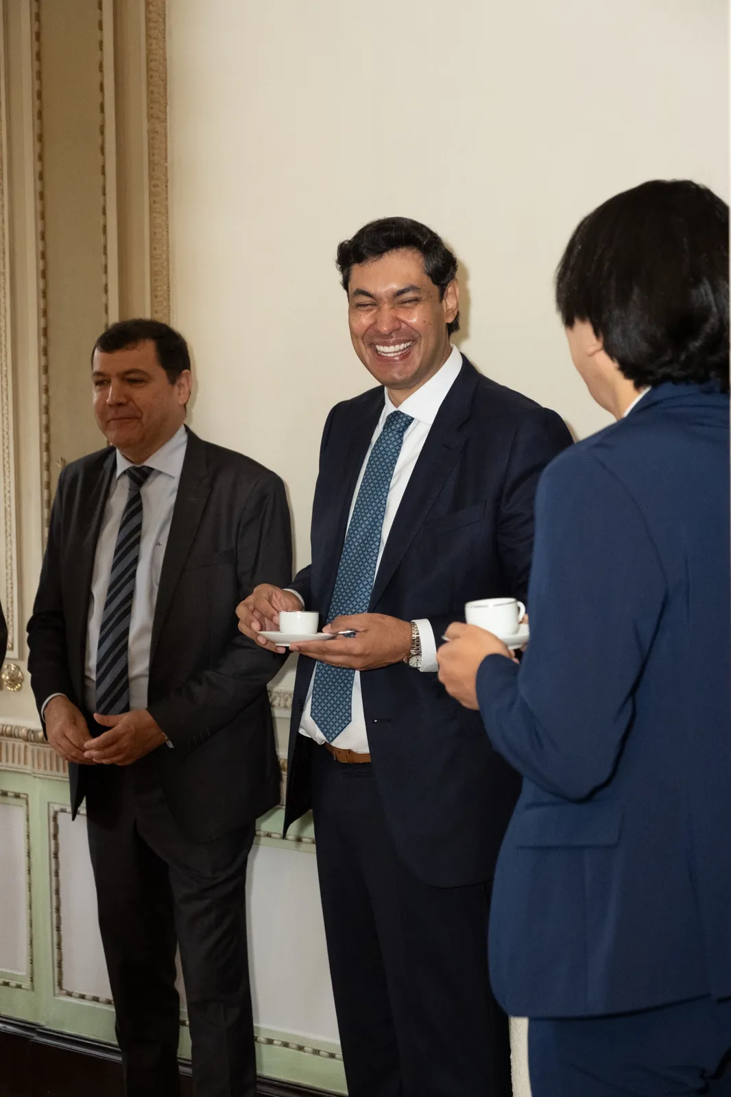 Três participantes de terno conversam de pé, com xícaras de café nas mãos, em salão de boiserie clara.