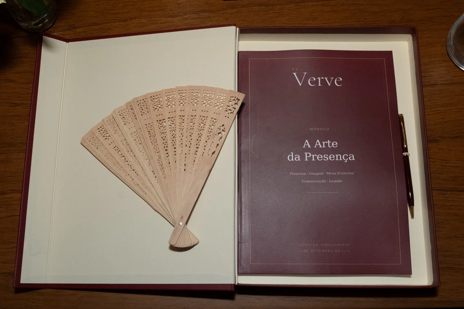 Estojo aberto com leque de madeira vazada à esquerda e o livro do participante do Módulo I à direita, ao lado de uma caneta.